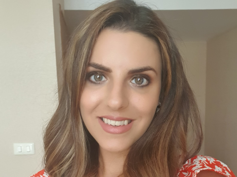 A woman with long, wavy hair and bright eyes smiles directly at the camera. She is wearing a floral-patterned blouse, and a softly lit background suggests a cozy indoor space.