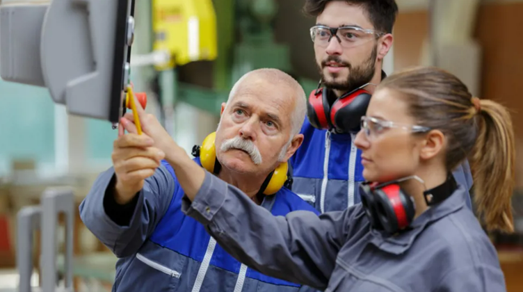 Two young workers wearing safety goggles and ear protection receive instruction from a senior worker in a workshop setting. The senior worker is pointing at a control panel while discussing with them, emphasizing a safety procedure. All three are dressed in gray work uniforms.