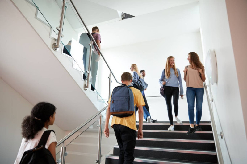 Students are walking up and down a modern staircase in a brightly lit space. Two girls are chatting on the stairs, while others are scattered around, some ascending and others descending. A boy with a backpack leads the way.