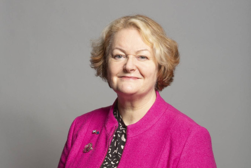 A woman with curly blonde hair, wearing a bright pink jacket over a patterned top, smiles confidently at the camera against a neutral gray background.