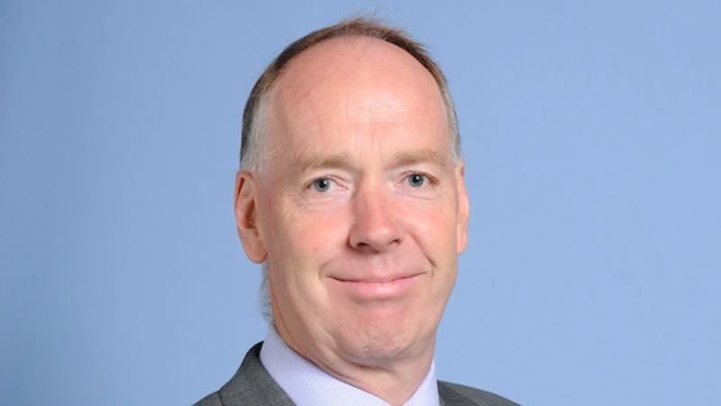 A man with short hair and a slight smile, wearing a grey suit and a light purple shirt, stands against a plain blue background.