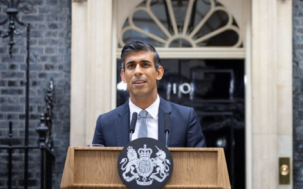 A man in a suit stands at a podium with the UK coat of arms, speaking outside a building with a large doorway and window.