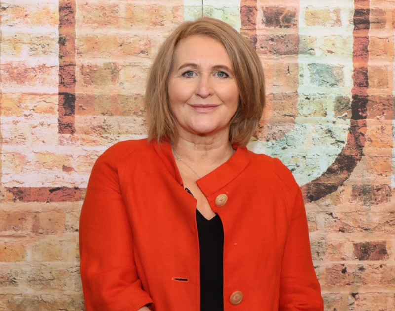 A woman with shoulder-length light brown hair smiles while wearing an orange blazer against a textured brick wall background.