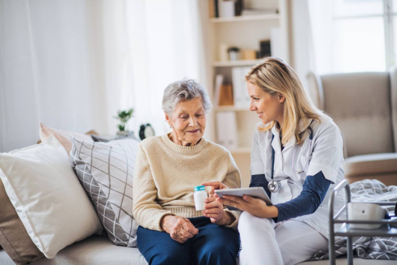 A healthcare professional is sitting next to an elderly woman in a cozy living room. The doctor, holding a tablet, discusses medication with the woman, who is holding a pill bottle. Both appear engaged and comfortable in the conversation.