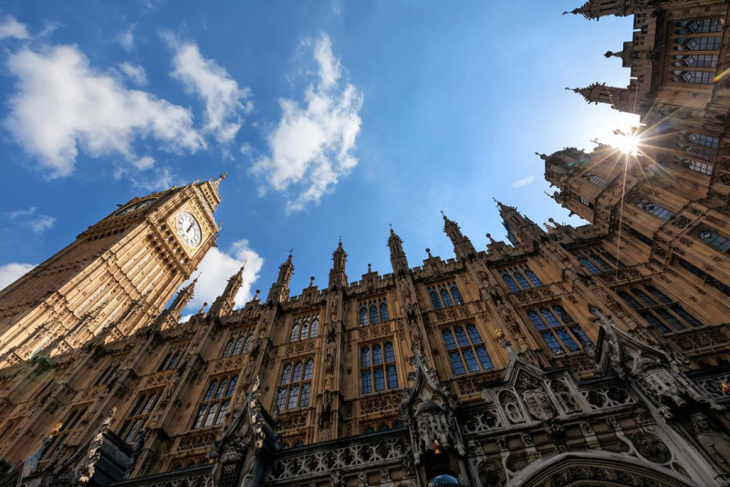 The iconic Big Ben clock tower rises above the ornate Victorian architecture of the Palace of Westminster, framed by a bright blue sky and scattered clouds. Sunlight glints off the tower's clock face.