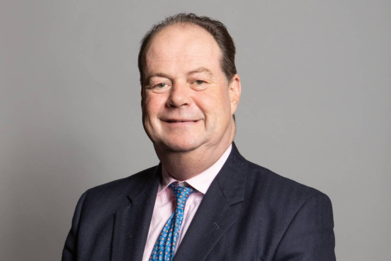 A smiling man in a dark suit and light pink shirt, wearing a patterned tie, stands against a plain gray background.