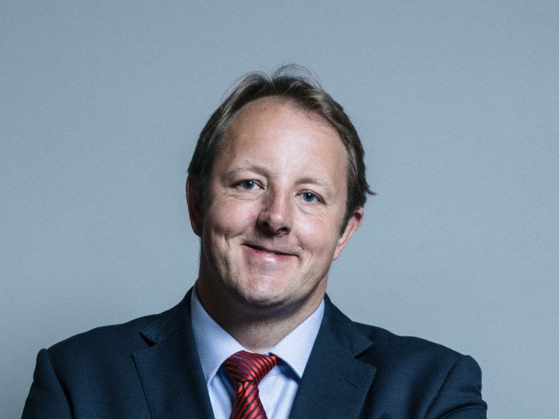 A man in a dark suit and red tie smiles confidently against a light gray background.