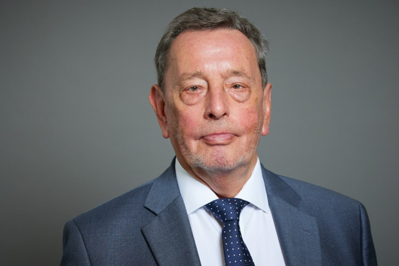 A middle-aged man with gray hair and a slight beard, wearing a dark blue suit and a white shirt with a patterned tie. He has a neutral expression and is facing the camera against a gray backdrop.