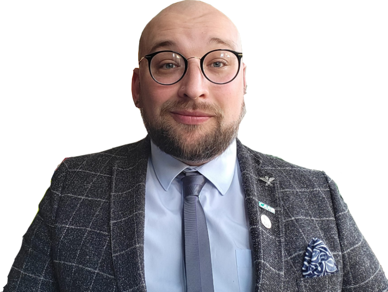 A man with a bald head and beard wears glasses, a gray checked suit, and a blue tie. He smiles warmly in front of a wall decorated with colorful hexagonal shapes and plants.