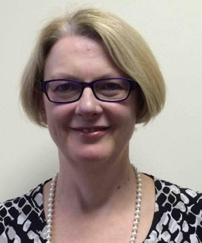 A smiling woman with short blonde hair and glasses, wearing a patterned top and a string of pearls.