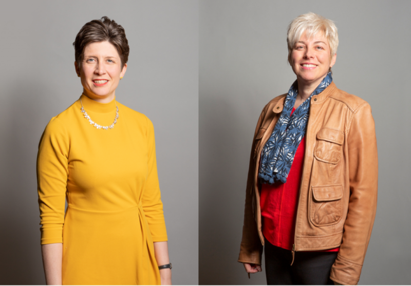 Two women standing side by side against a gray background. The woman on the left wears a bright yellow dress with a necklace, smiling confidently. The woman on the right is dressed in a red top with a patterned scarf and a brown leather jacket, also smiling.