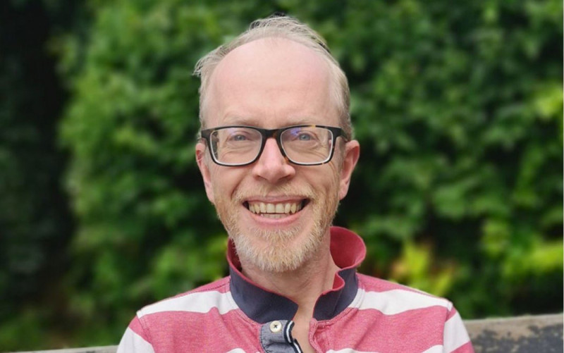 A smiling man with glasses and short, light-colored hair wears a red and white striped shirt. He is seated outdoors with lush greenery in the background.