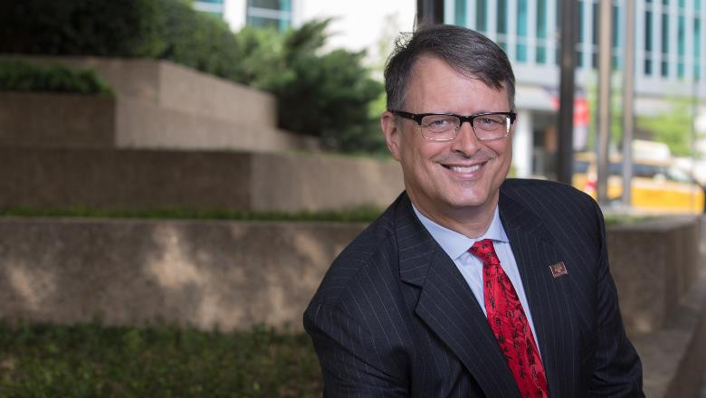 A smiling man with glasses wearing a black pinstriped suit and a red tie poses outdoors, with green shrubbery and a modern building in the background.