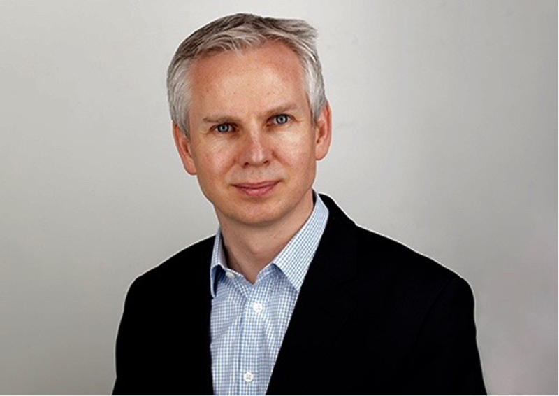 A man with silver hair wearing a dark blazer over a light blue checked shirt poses for a formal portrait against a neutral background.