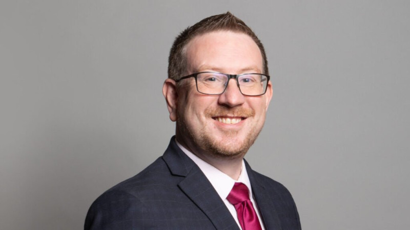 A man with short, brown hair and glasses smiles wearing a blue suit and a pink tie against a light gray background.