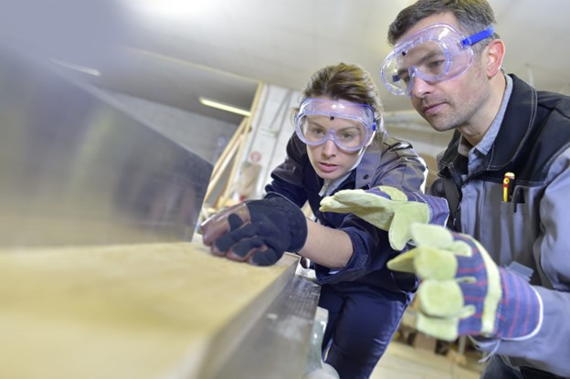 A woman and a man wearing safety goggles and gloves are carefully examining a piece of wood in a workshop. The woman is focused on measuring, while the man is guiding her.