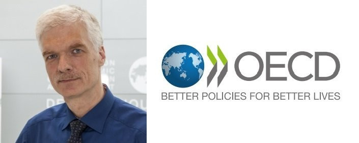 A man with gray hair and a mustache is wearing a blue shirt, standing beside the OECD logo, which features a globe and the tagline "Better policies for better lives."