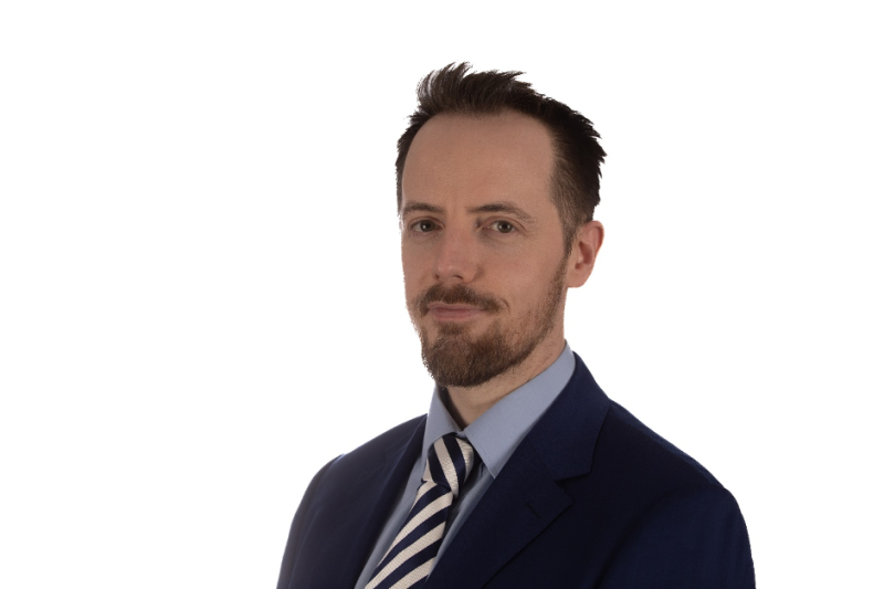 A man with a short beard and well-groomed hair is wearing a navy blue suit and a light blue shirt with a striped tie. He is looking confidently towards the camera with a neutral expression against a plain white background.