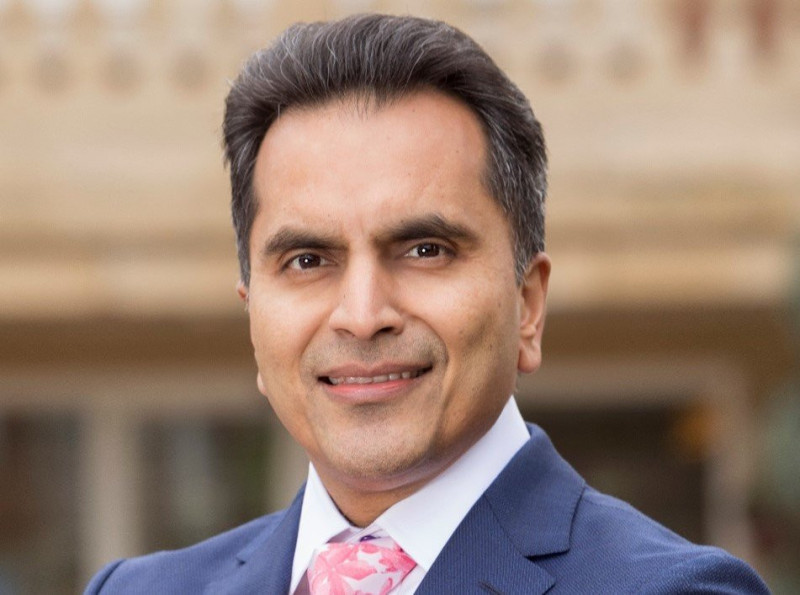 A smiling man in a dark suit and pink tie stands outdoors, with a soft-focus background featuring warm, neutral tones.