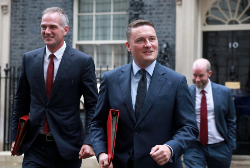 Three men in business attire walk together outside a building. Two men are smiling and carrying folders, while the third man follows closely behind. The setting appears to be a formal office environment.