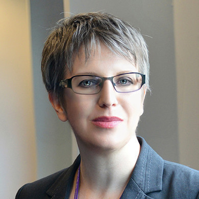 A woman with short gray hair and glasses, wearing a dark blazer, looks directly at the camera with a slight smile. She appears professional and confident.