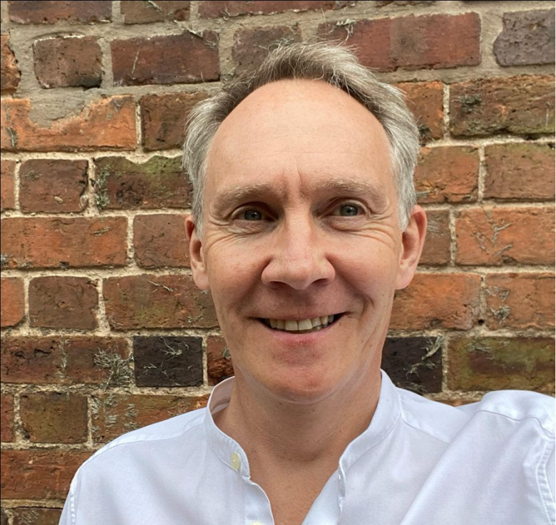 A smiling person with short gray hair wearing a white shirt stands in front of a rustic brick wall.