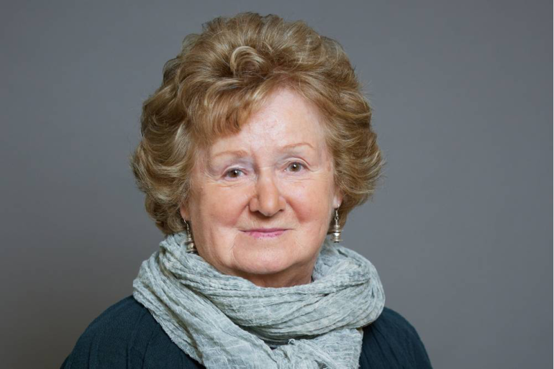 An elderly woman with curly light brown hair smiles warmly at the camera, wearing a light gray scarf over a dark top. A neutral gray background adds a soft touch to the portrait.