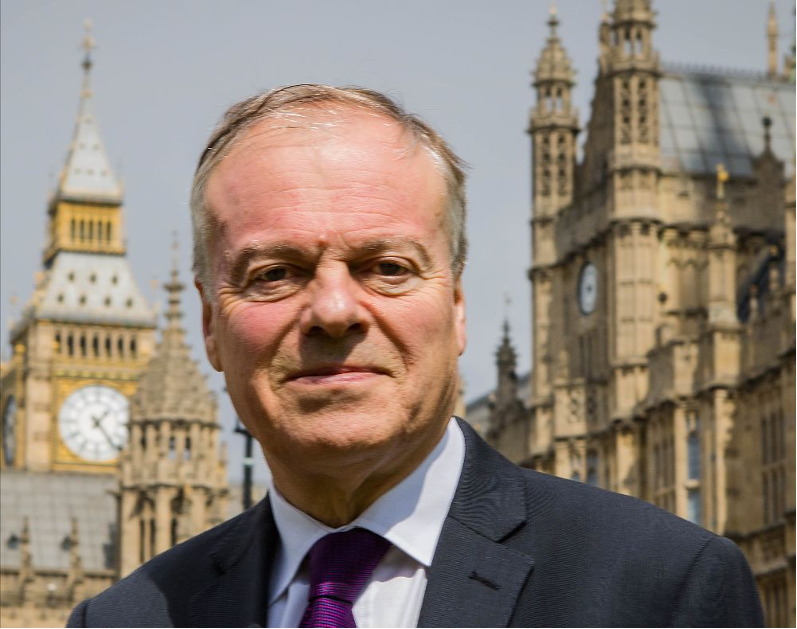 A man in a dark suit and purple tie stands in front of the Houses of Parliament in London, with Big Ben visible in the background. The sky is clear, and the scene conveys a formal atmosphere.