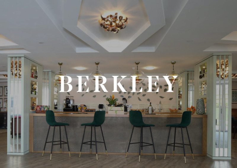 Stylish interior of a bar with a sleek design, featuring high stools, decorative lighting fixtures, and a floral centerpiece. The name "BERKLEY" is prominently displayed in a bold font.