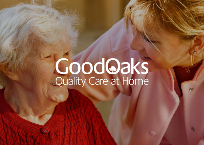 A caregiver interacts warmly with an elderly woman, both smiling and engaged in conversation. The background features soft lighting, and the text "GoodOaks Quality Care at Home" is prominently displayed.