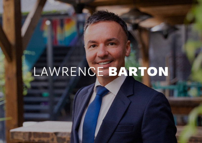 A professional man with short dark hair and a suit smiles confidently in a vibrant outdoor setting, with stairs and colorful artwork in the background.