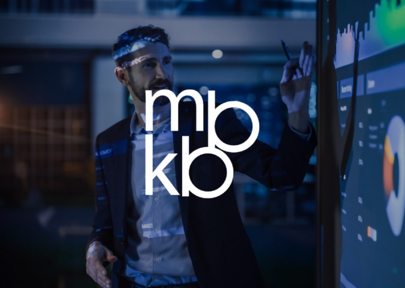 A man in a suit gestures towards a digital screen displaying charts and graphs, with a blue light reflecting on his face. The letters "mbkb" are prominently displayed in the foreground.