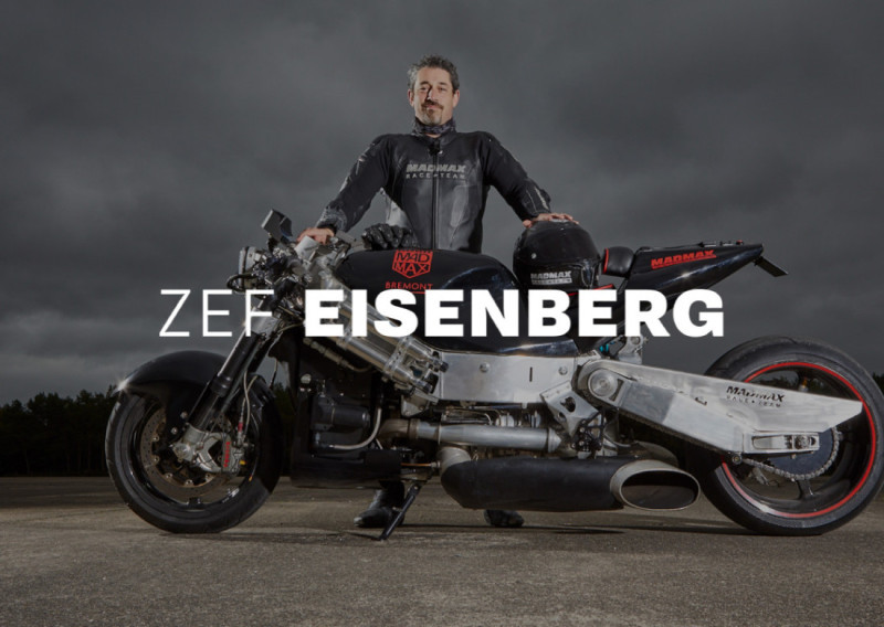 A man stands beside a futuristic motorcycle, wearing a black racing suit and helmet. He appears confident with a serious expression, set against a cloudy sky. The text "Zef Eisenberg" is prominently displayed across the image.