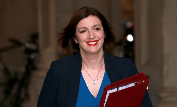 A woman with medium-length brown hair and bright red lipstick smiles while holding a red folder. She wears a blue blouse and a dark blazer, walking through an architectural setting.