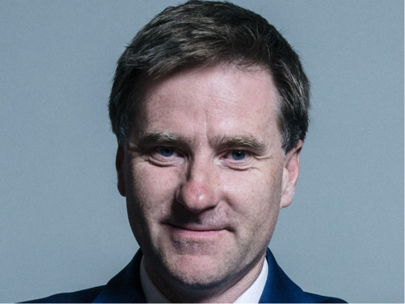 A man with short brown hair and blue eyes is smiling against a gray background. He is wearing a dark suit and tie.