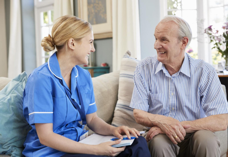 A healthcare worker in a blue uniform engages in a friendly conversation with an older man sitting on a couch. They both smile, suggesting a warm and supportive interaction. The setting is bright and comfortable, with soft furnishings and natural light.