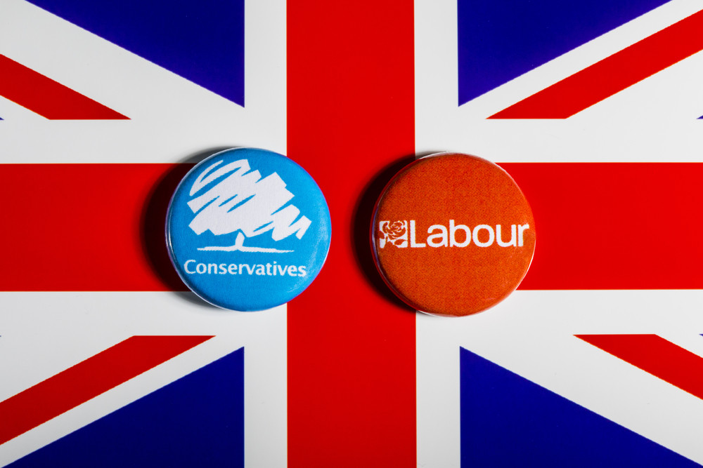 Two political buttons placed on a background featuring the Union Jack. One button is blue with the word "Conservatives" and a stylized graphic, while the other is orange with the word "Labour."