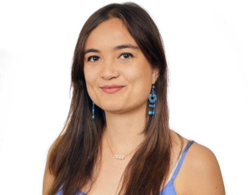 A smiling young woman with long, dark hair wearing a blue dress and turquoise earrings, set against a white background.