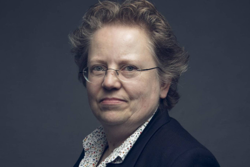 A middle-aged woman with short, curly hair and glasses smiles softly, wearing a dark blazer over a light, patterned shirt. The background is a muted gray.