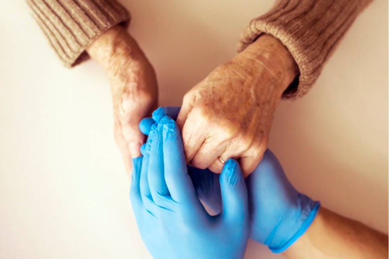 Two hands are holding each other, one showing age with wrinkled skin and the other wearing a blue medical glove. The warm gesture suggests compassion and care.