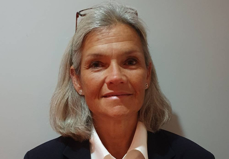 A woman with shoulder-length gray hair smiles warmly, wearing a dark blazer over a white shirt. She has earrings and glasses resting on her head. The background is a plain light color.