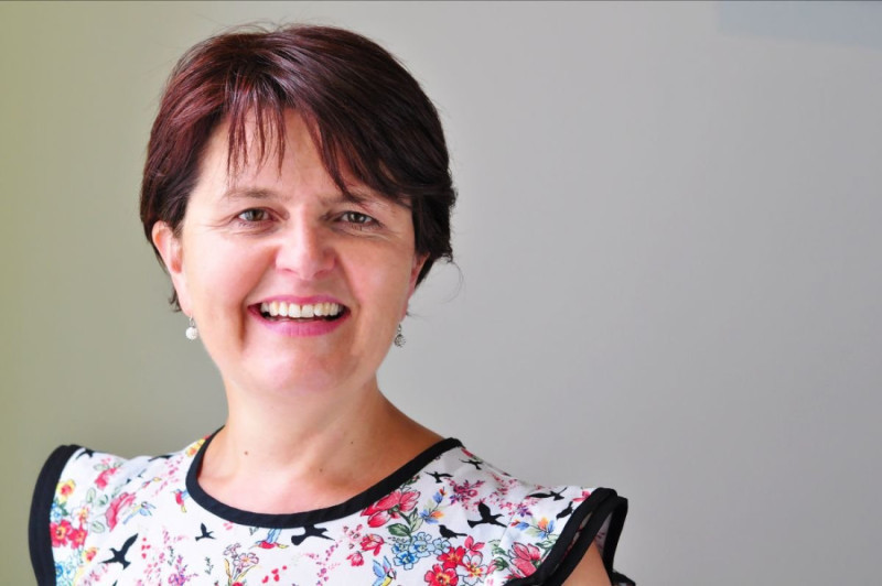 A smiling woman with short, dark hair wearing a floral blouse stands against a light background.