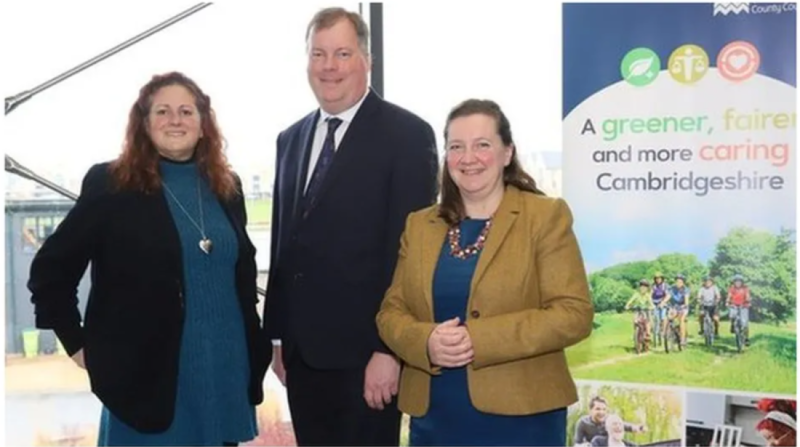 Three professionals stand together, smiling in front of a poster that promotes a greener and fairer Cambridgeshire. The individuals are dressed in smart attire, with a view of a cityscape visible through a window behind them.