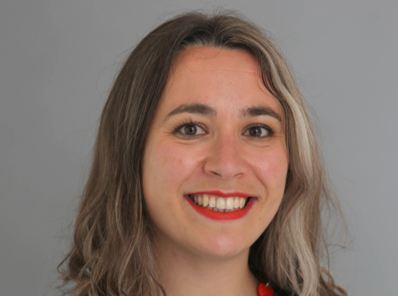 A smiling person with shoulder-length wavy hair and red lipstick, set against a neutral gray background.