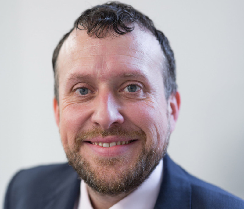 A smiling man with curly hair and a beard, wearing a suit jacket and pink shirt, poses for the camera against a light background.