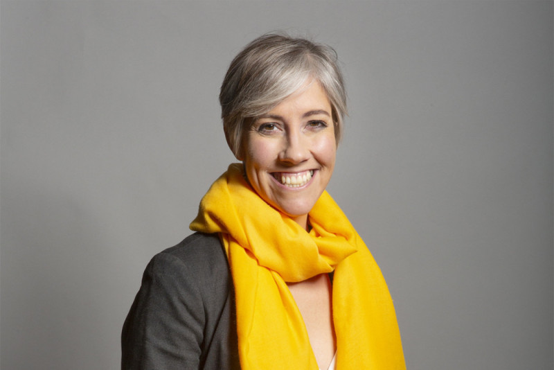 A woman with short, silver hair smiles warmly while wearing a bright yellow scarf and a gray blazer against a neutral background.