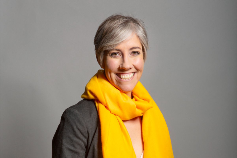 A woman with short, silvery hair smiles warmly while wearing a bright yellow scarf and a dark blazer. The background is light gray.