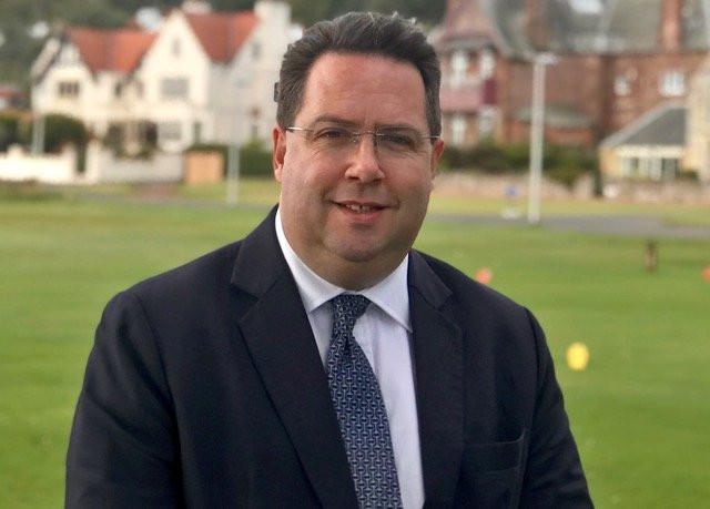 A man in a dark suit and patterned tie standing on a green field with buildings in the background.