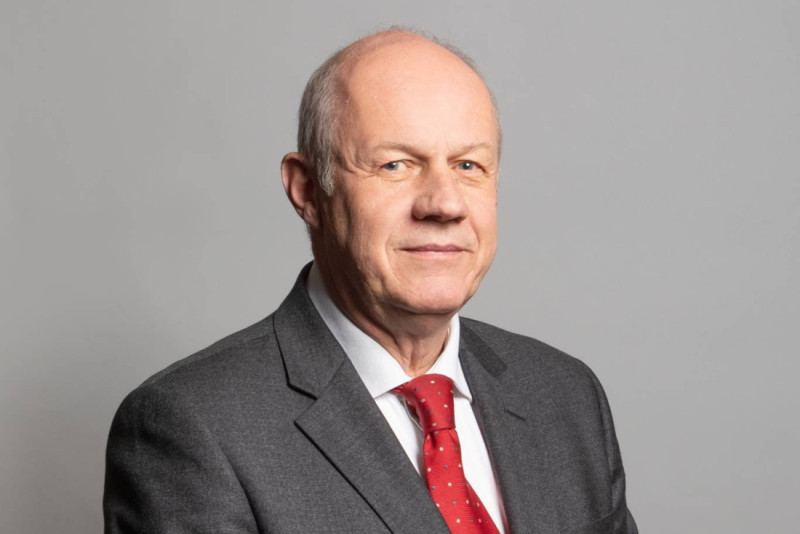 A middle-aged man in a suit with a red tie poses against a neutral gray background. He has short, graying hair and is smiling slightly.