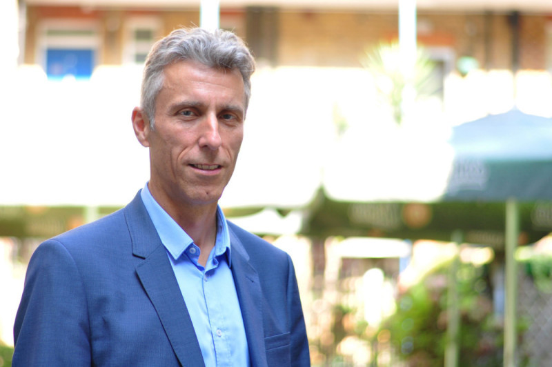 A man with short gray hair stands outdoors, wearing a blue suit and light blue shirt. He appears confident, with a blurred background of greenery and outdoor seating.